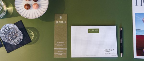 Teller mit Pralinen, Glas, Notizblock, Stift und Broschüre auf grünem Tisch