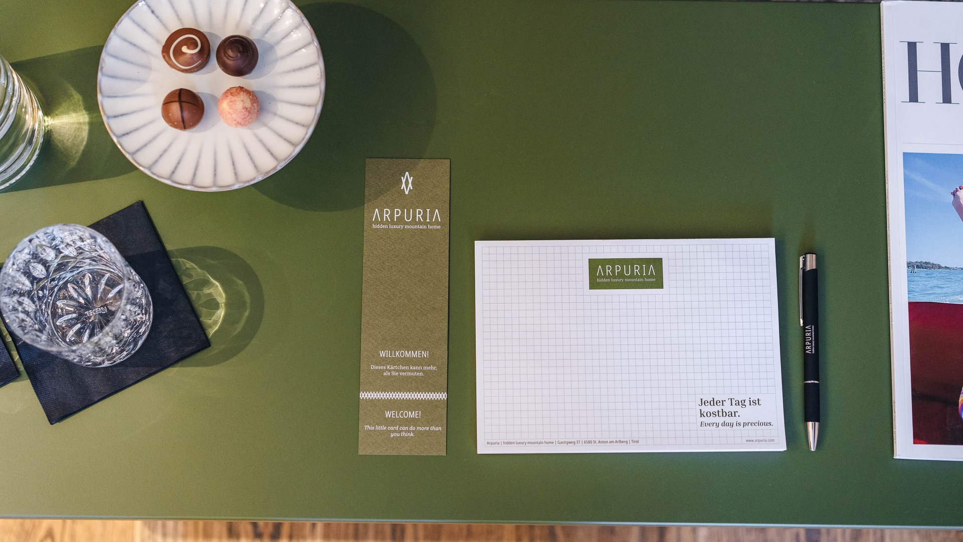 Teller mit Pralinen, Glas, Notizblock, Stift und Broschüre auf grünem Tisch