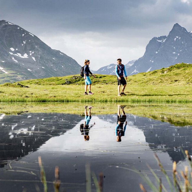 Ihr Urlaub am Arlberg im Sommer – das Wanderparadies Zwei Wanderer auf einer grünen Wiese mit Bergreflexionen im Wasser