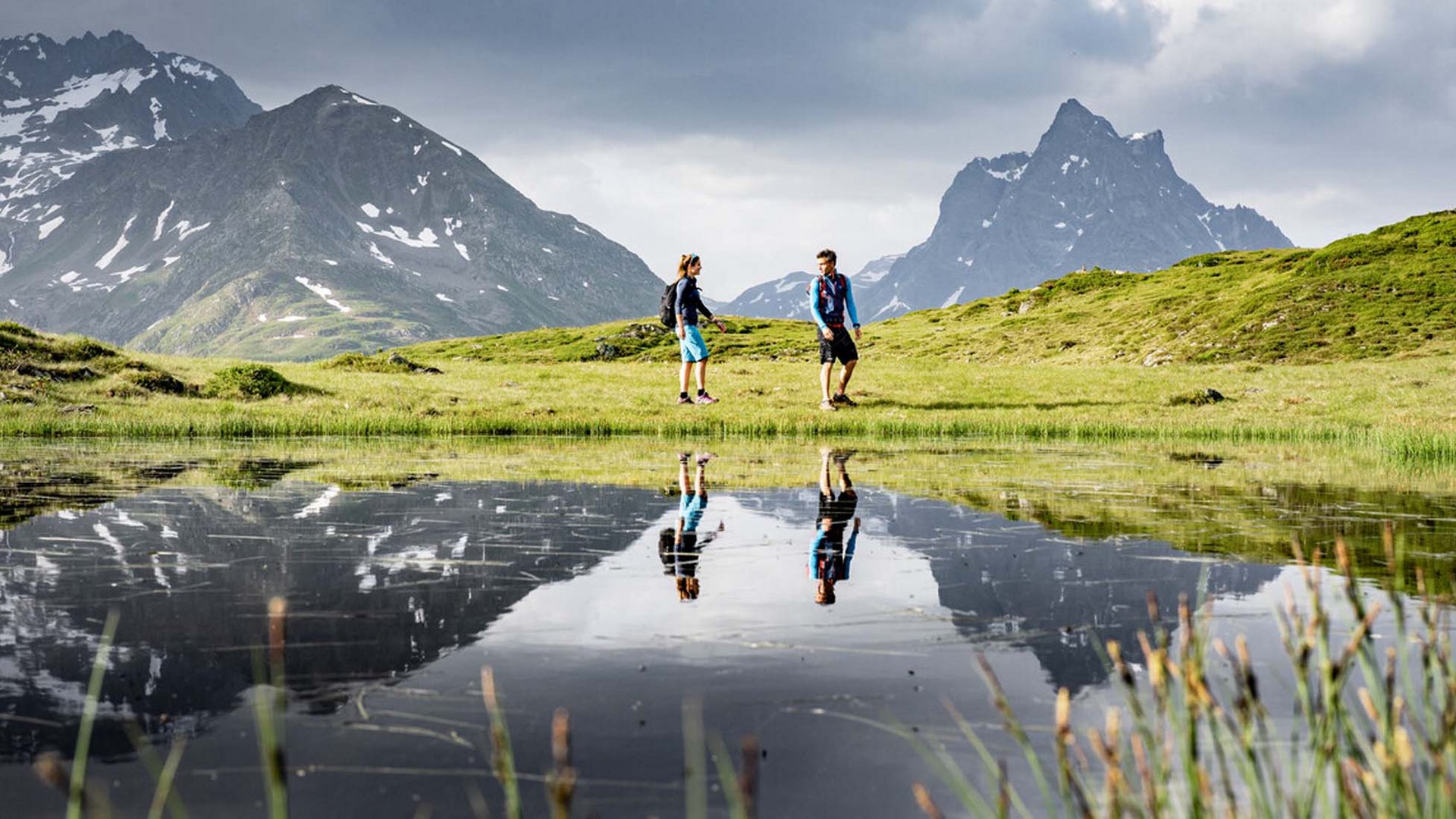 Two hikers on green meadow with mountain reflections in water