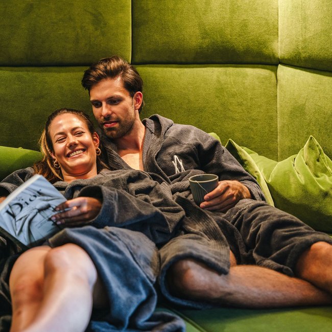 Pärchen entspannt auf grünem Sofa in Bademänteln beim Lesen und Tee trinken