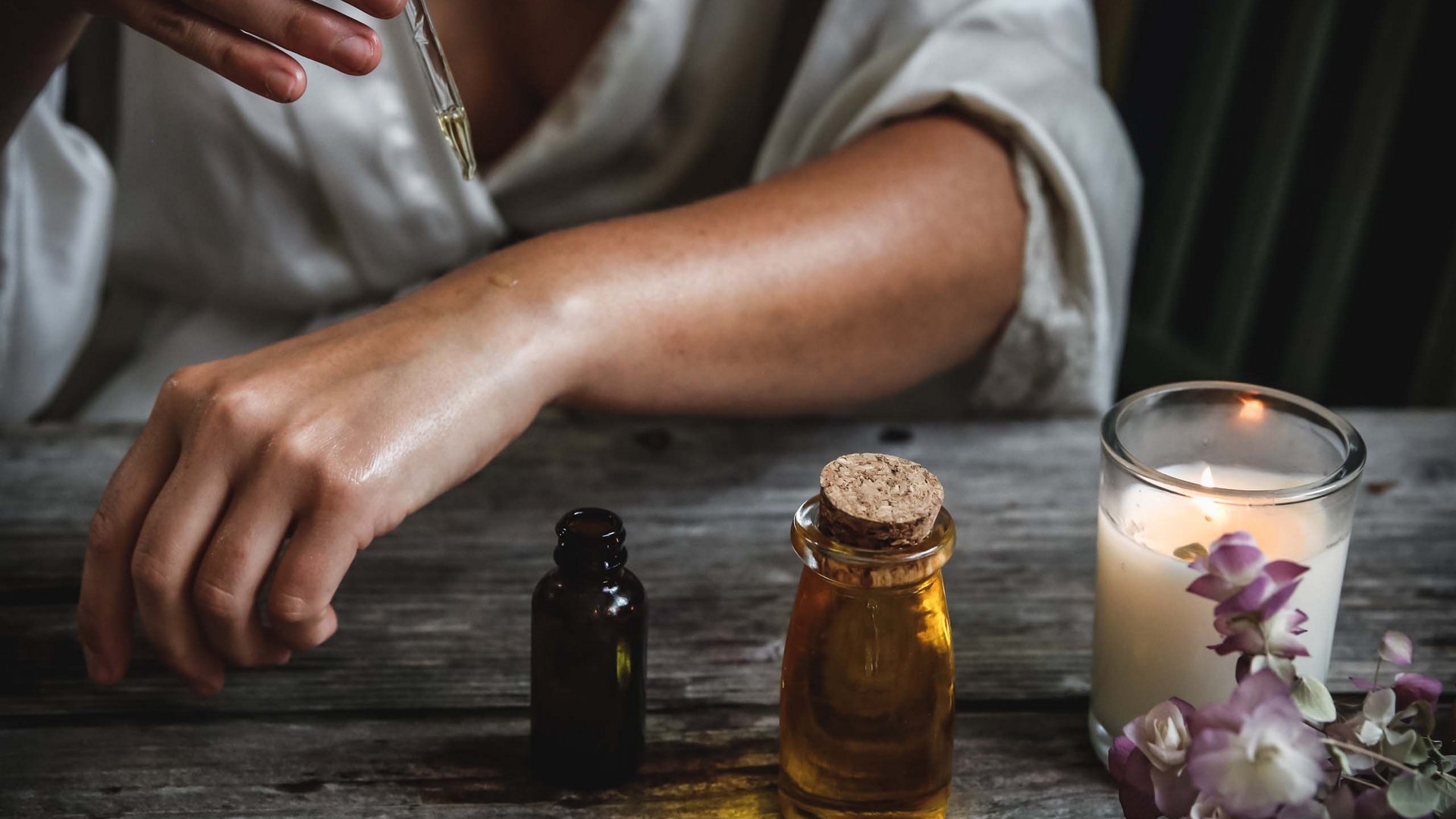 Person applying essential oil on arm with dropper beside scented candle and flowers