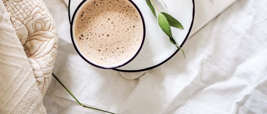 Cup of coffee on saucer with green leaves and white flowers on bedspread
