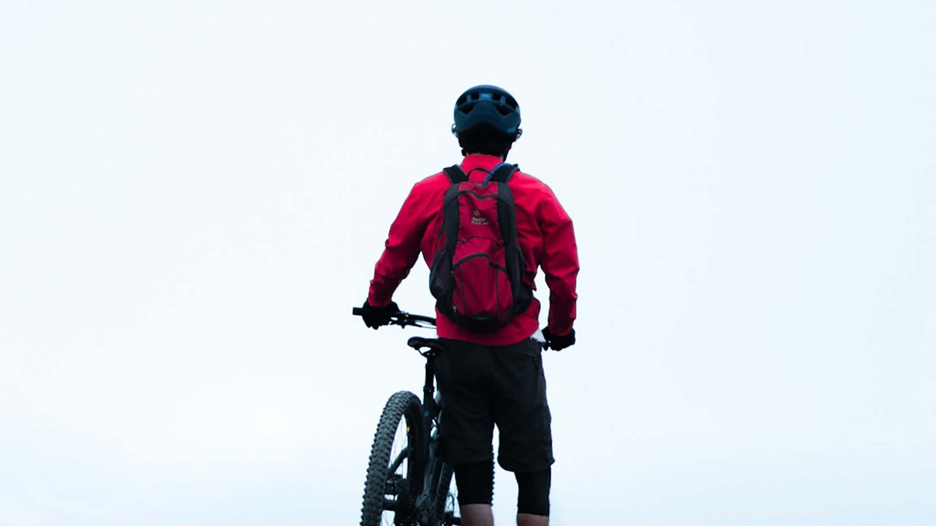 Mountain biker standing with bike on grassy hill against bright sky