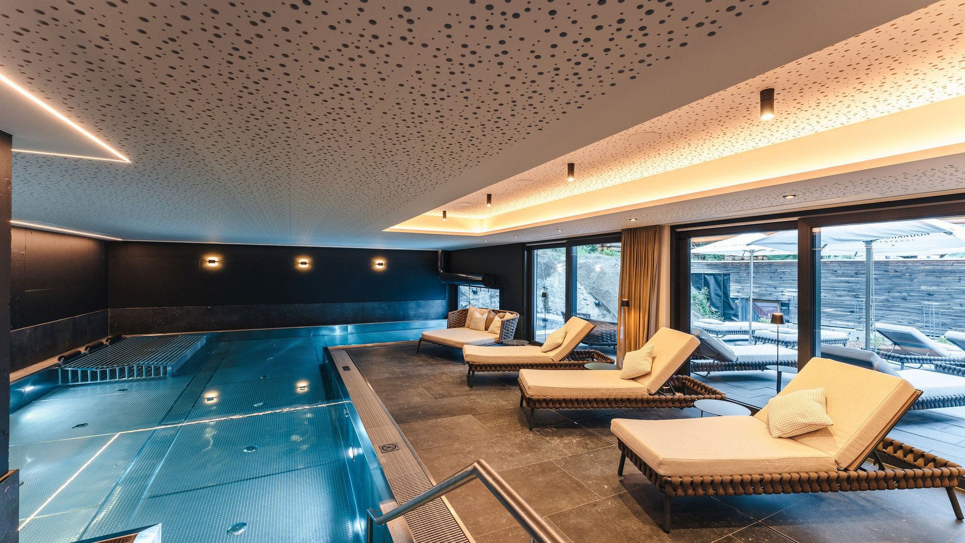 Modern indoor pool with lounge chairs and large windows opening to a terrace