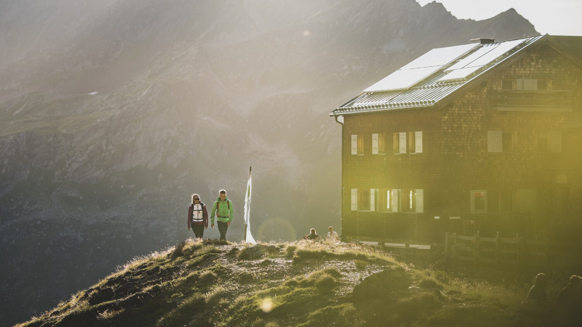 Hikers near a mountain hut with sunlight and mountains in the background