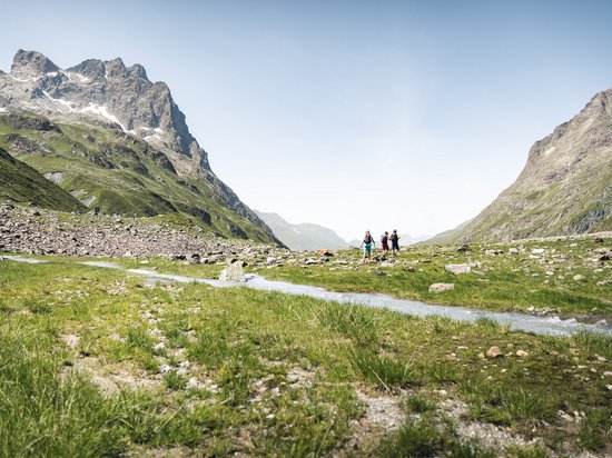 St. Anton: Sommerurlaub, Winterspaß Drei Wanderer in einem grünen Tal mit Bergen und Bach unter blauem Himmel