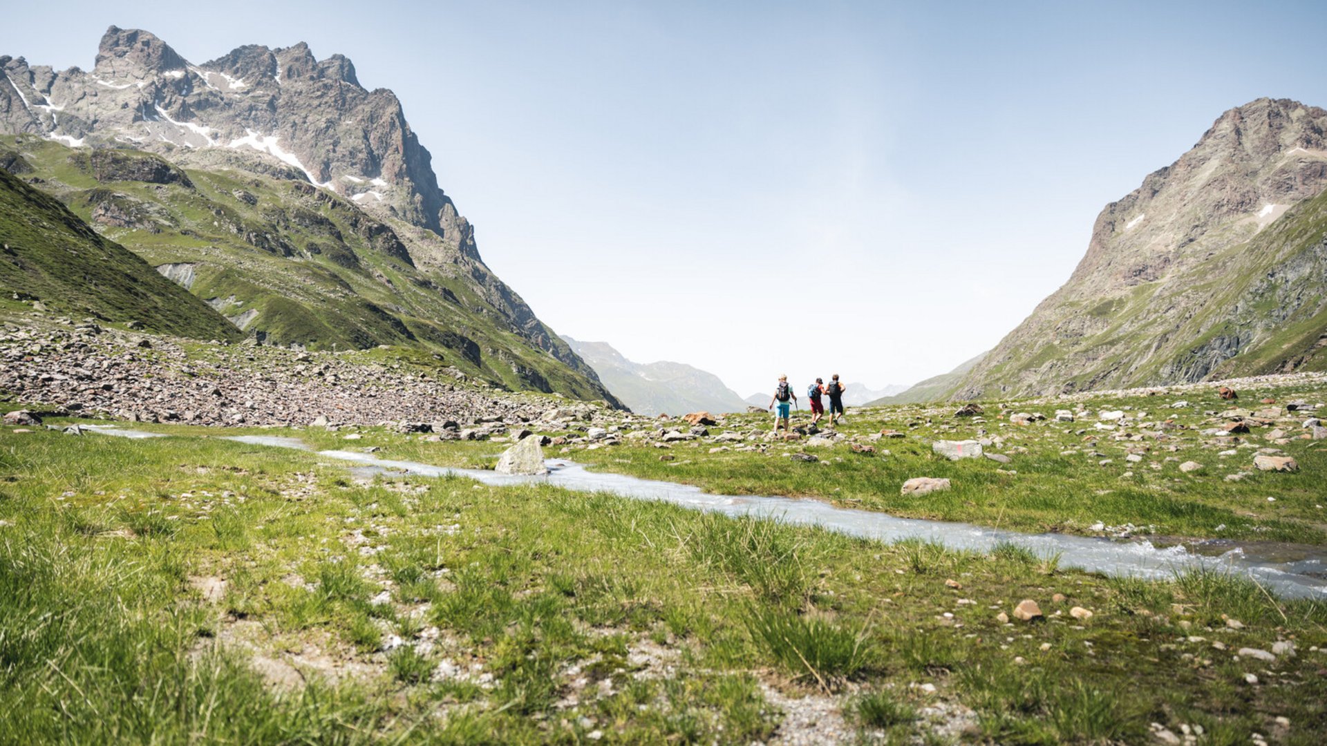 Drei Wanderer in einem grünen Tal mit Bergen und Bach unter blauem Himmel