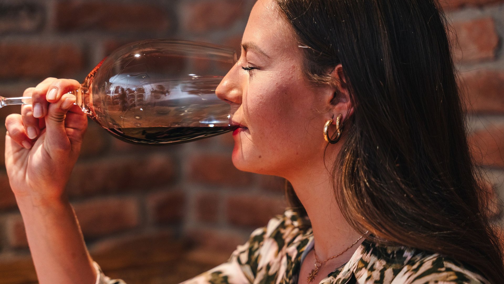 Woman drinking red wine from a glass in front of a brick wall