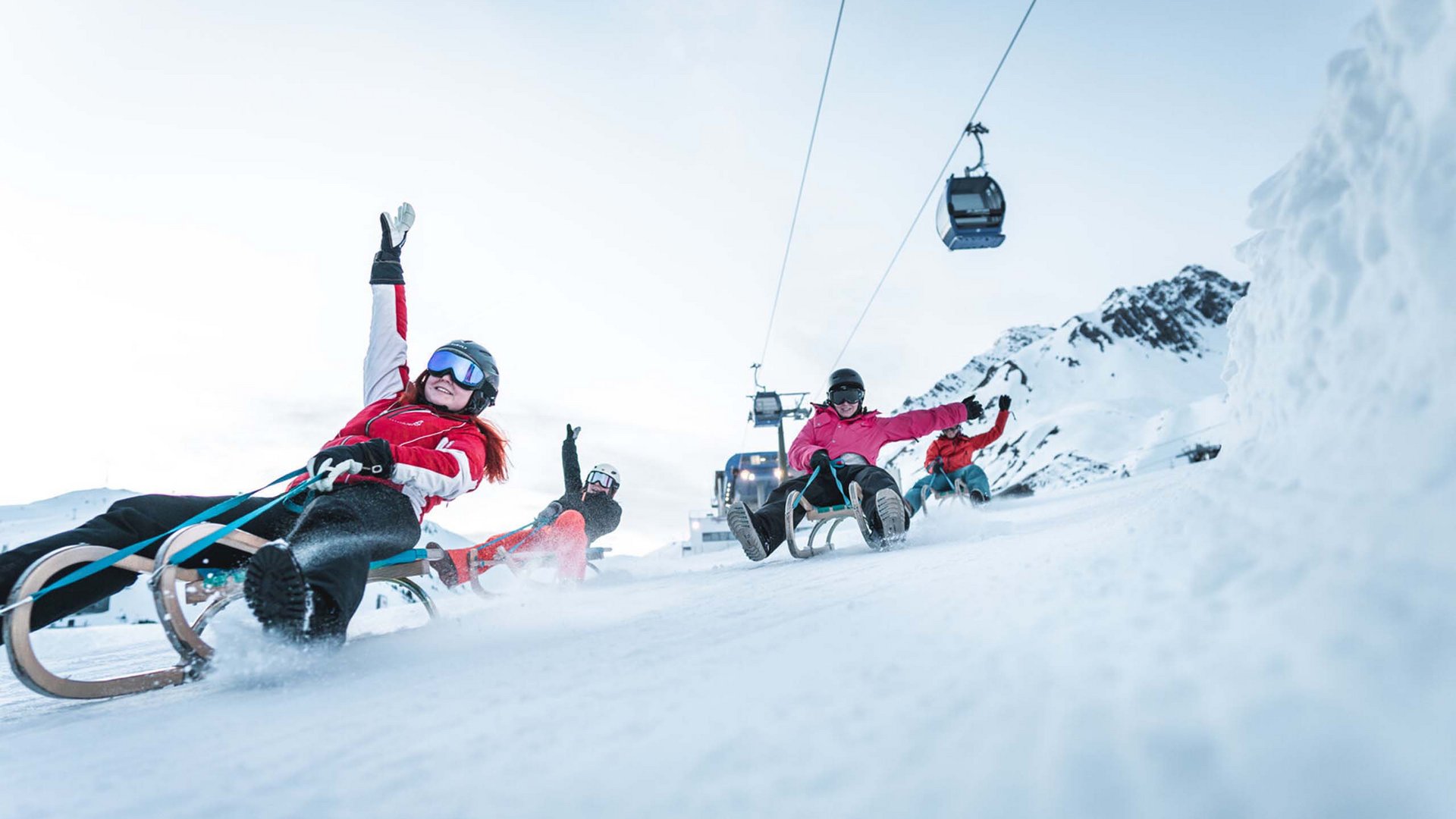 Four people sledding down a snowy hill with a cable car in the background