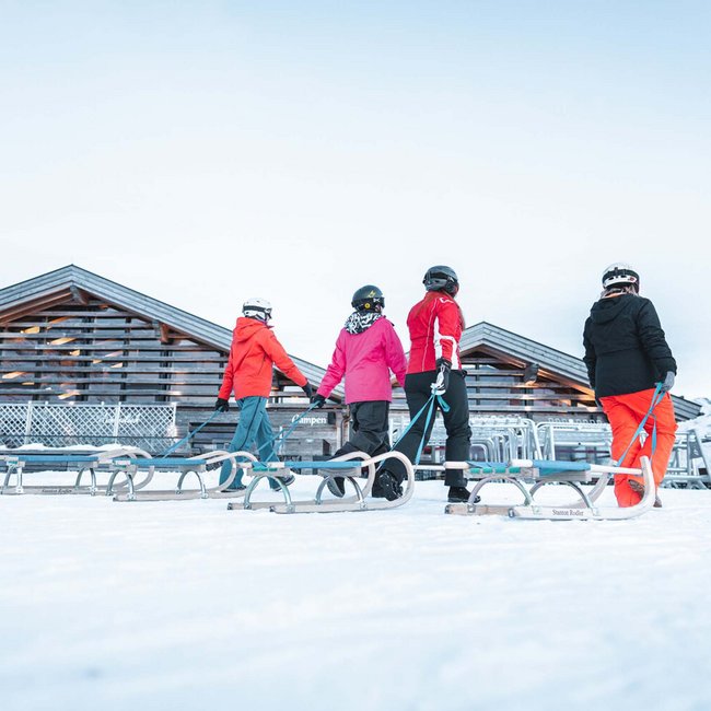 Four people in snow with sleds in front of wooden cabin and mountains