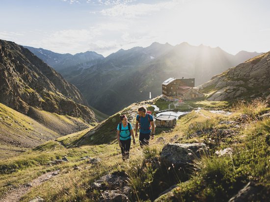 St. Anton: Sommerurlaub, Winterspaß Zwei Wanderer steigen auf einem Bergweg bei Sonnenschein zu einer Berghütte auf