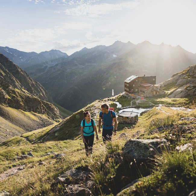 Zwei Wanderer steigen auf einem Bergweg bei Sonnenschein zu einer Berghütte auf