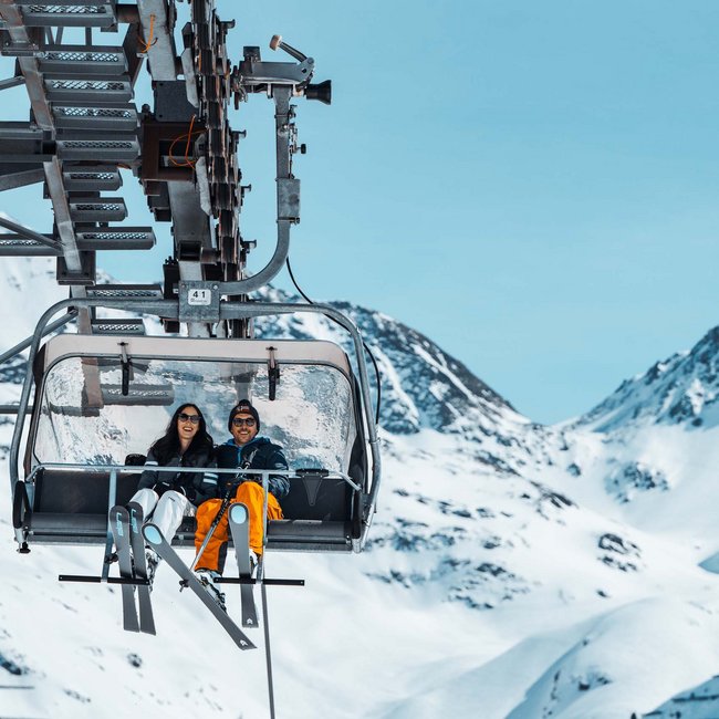 Zwei Skifahrer auf Sessellift vor schneebedeckten Bergen bei klarem Himmel