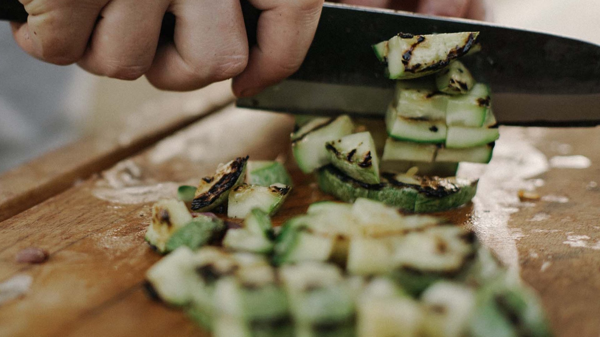 Person chopping grilled zucchini on a wooden cutting board with a knife