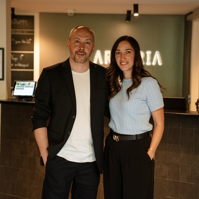Willkommen im Arpuria. Ein Mann und eine Frau stehen vor einer Rezeption und lächeln in die Kamera