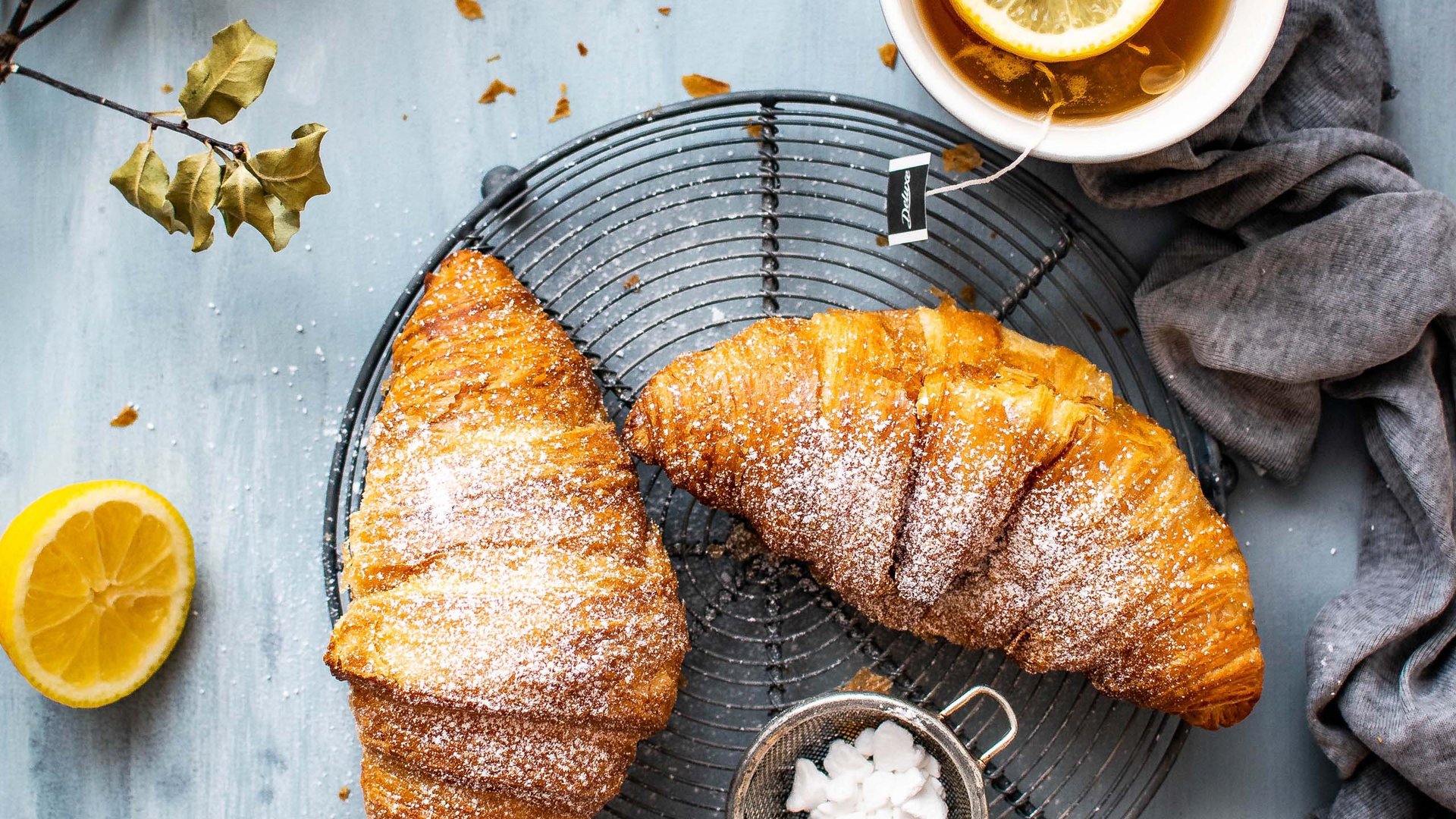 Two croissants with powdered sugar, lemon tea with lemon slice on blue surface
