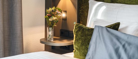 Schlafzimmer mit grünem Kopfteil, Kissen und Nachttisch mit Vase und Blumen