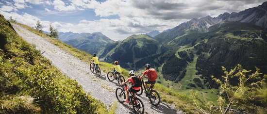 Familie radelt auf Bergweg mit Alpenpanorama unter bewölktem Himmel